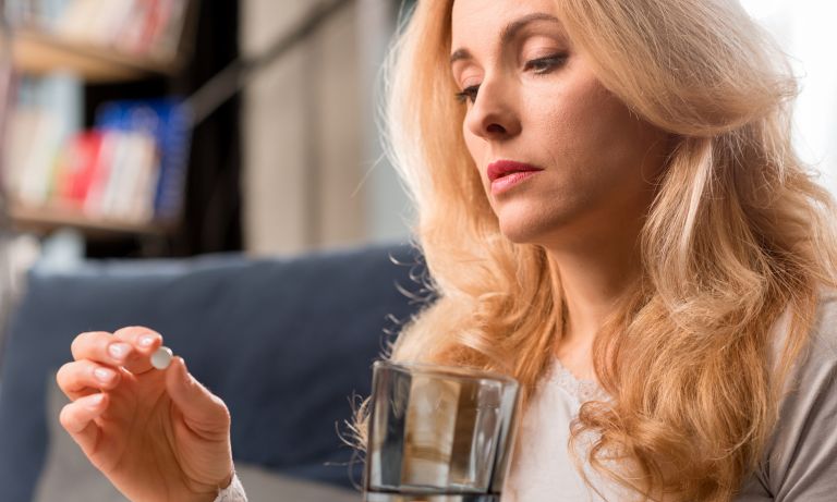 Woman holds pill between thumb and forefinger, contemplating it, glass of water in other hand